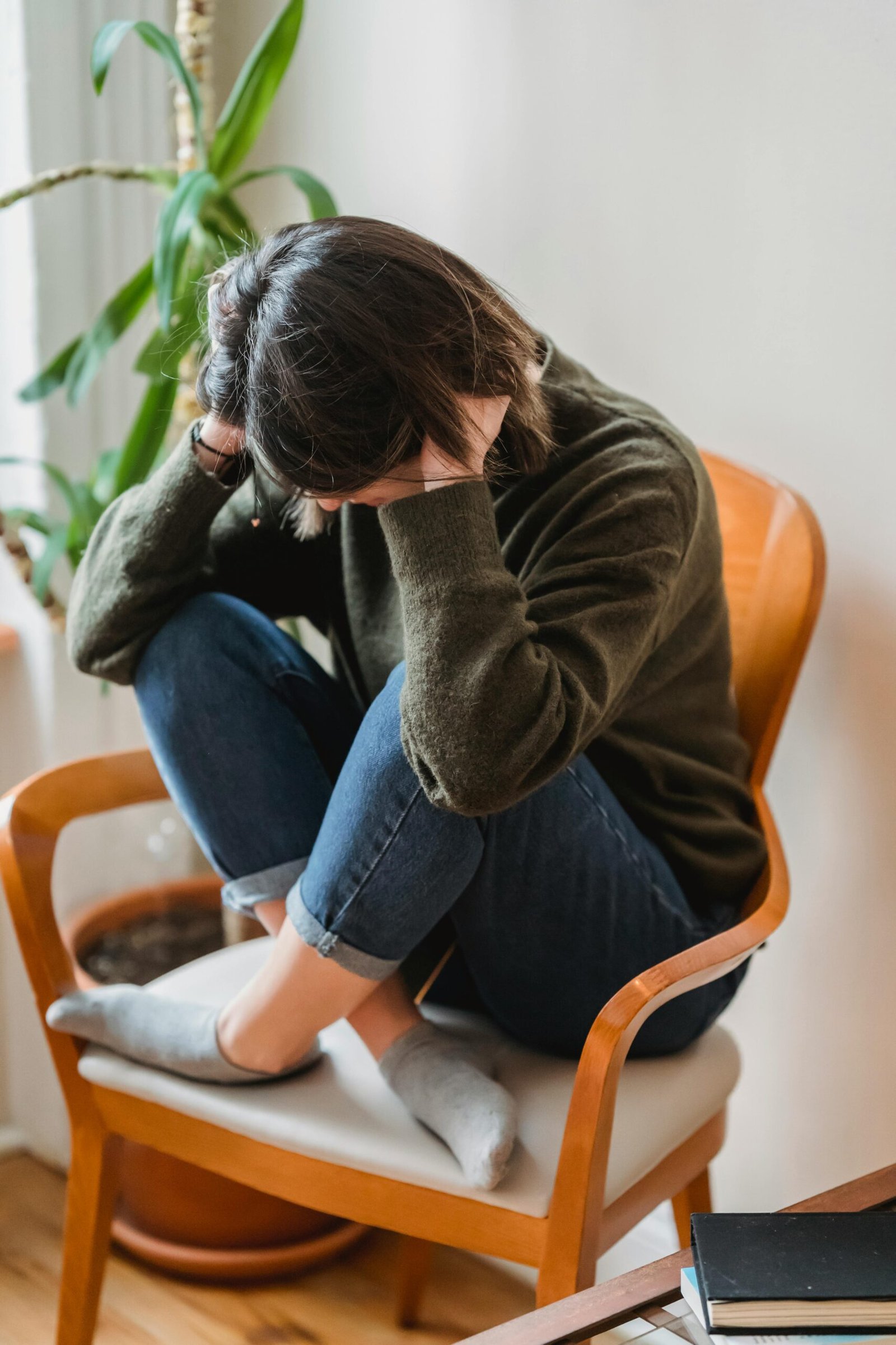 Full body of unrecognizable young worried female in casual clothes sitting on chair with crossed legs and covering ears with hands at home